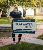 Assistant Vice President, Morgan Fornoff standing in front of house on the sidewalk behind a sign that says, "Financing Provided by: Flatwater Bank"