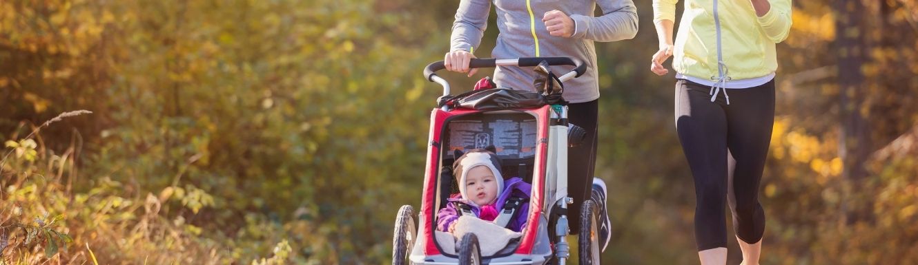 A dad pushing his infant daughter in a jogging stroller in an outdoor park on a fall morning. The mom is jogging right next to them.