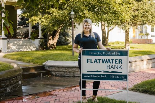 Assistant Vice President, Morgan Fornoff, standing in front of house with a sign that says, "Financing provided by Flatwater Bank"
