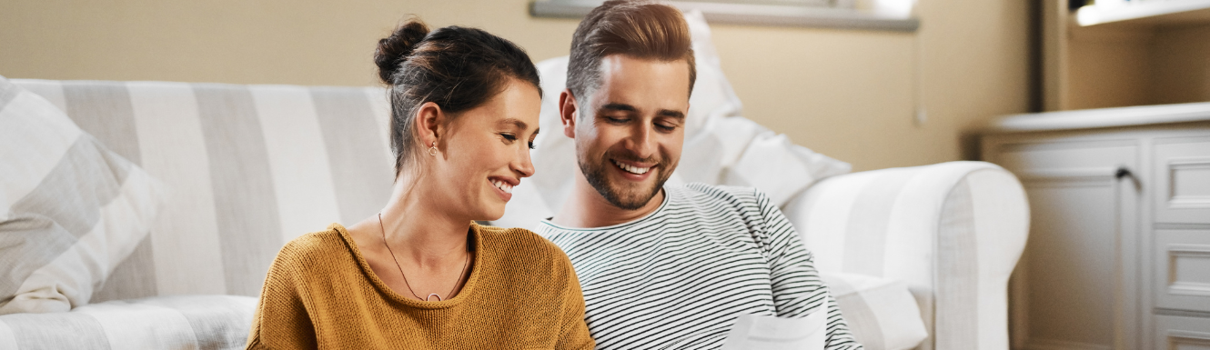 younger couple smiling and sitting in front of their couch looking at financial paperwork