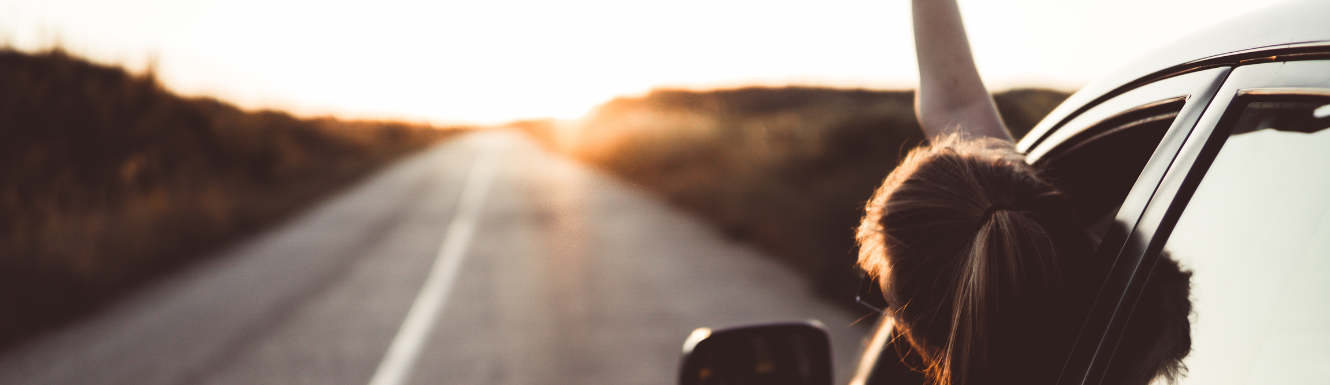 person leaning out of car window while going down a long paved road with sun shining